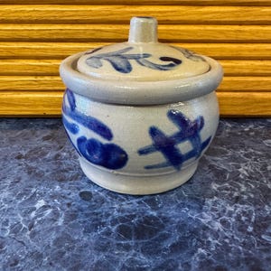 May include: A small, round ceramic jar with a lid. The jar is off-white with blue floral designs and a matching lid. The lid has a small handle. The jar is sitting on a blue and gray surface.