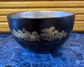 May include: A black bowl with a silver interior and a decorative gold and red landscape design around the exterior. The bowl sits on a blue marble-patterned surface.