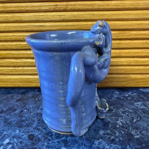 May include: A handmade blue ceramic mug with a lizard-shaped handle. The mug has a cylindrical shape and a textured surface. The lizard is detailed with a curled tail and a head resting on the rim. The mug is a unique piece of pottery.