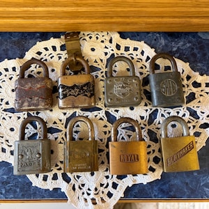 May include: A collection of eight antique padlocks in various sizes and finishes, arranged on a white lace doily over a blue surface. The locks display signs of age, with some showing rust and wear. The locks have different brand names.