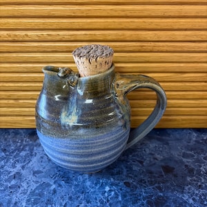 May include: A round ceramic pitcher with a glazed finish in shades of dark blue and brown, featuring a ribbed texture on the lower body, a single handle on the side, and a natural cork stopper inserted into the top opening.