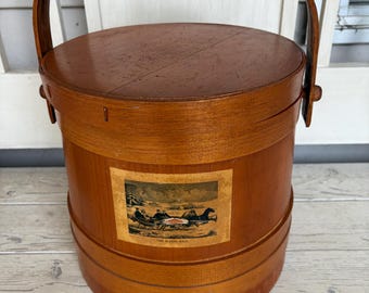 May include: A cylindrical wooden bucket with a swing handle and a fitted lid. The exterior features a rectangular print depicting a winter horse-drawn sleigh scene with the text "THE SLEIGH RACE" printed in small black letters at the bottom.
