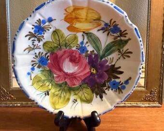 May include: A round, scalloped-edge ceramic plate featuring a hand-painted floral design. The center displays a large pink rose, a purple flower, and a yellow tulip surrounded by green leaves and small blue blossoms. The rim has blue painted accents.