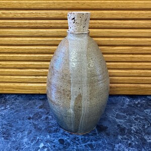May include: A handmade, ceramic bottle with a cork stopper. The bottle has a rounded shape with a textured, speckled surface in shades of tan and gray. A vertical, lighter-colored stripe runs down the front.