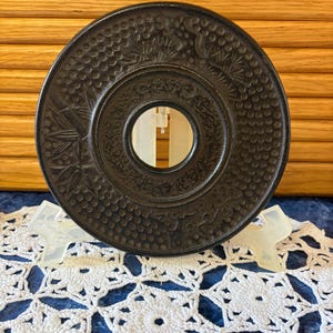 May include: A circular black cast iron trivet featuring a central hole and an embossed pattern of bamboo stalks and small dots. The object is supported by a clear plastic stand, showcasing the detailed relief work on its dark, textured surface.