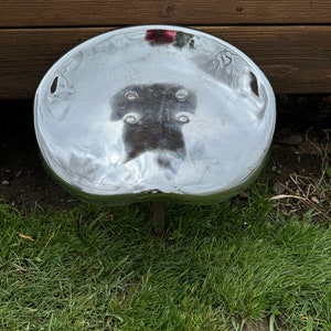 May include: A silver metal stool with a round seat and a single leg. The stool is sitting on a patch of green grass.