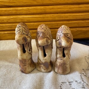 May include: Three ceramic figurines with a brown and white glaze. Each figurine has a unique face and a hole in the front. The figurines are standing on a white crocheted surface.