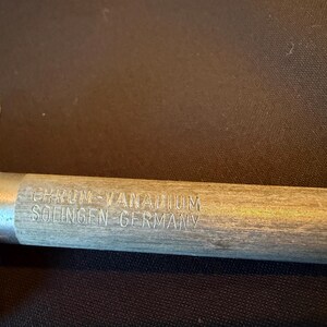May include: A metal sharpening rod with a wooden handle featuring the engraved text CHROM-VANADIUM SOLINGEN-GERMANY. The tool has a metallic, cylindrical shaft and a rounded, polished metal end cap near the handle, showing a clear, detailed view.