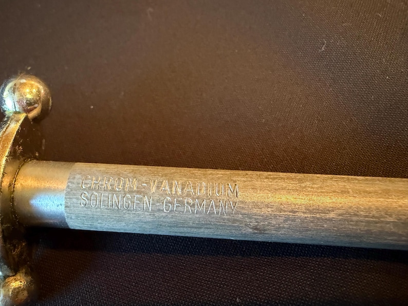 May include: A metal sharpening rod with a wooden handle featuring the engraved text CHROM-VANADIUM SOLINGEN-GERMANY. The tool has a metallic, cylindrical shaft and a rounded, polished metal end cap near the handle, showing a clear, detailed view.
