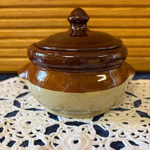 May include: A small, round ceramic pot with a lid, featuring a two-tone glaze. The upper portion and lid are a glossy dark brown, while the lower body is a lighter tan color. The pot includes two small side handles and a knob on the lid.