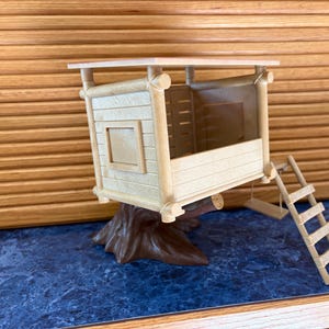 May include: A miniature wooden treehouse with a brown tree trunk base, a small ladder, and a swing. The treehouse has a light brown exterior with a square window and a flat roof. The background is a blue surface and a wooden wall.