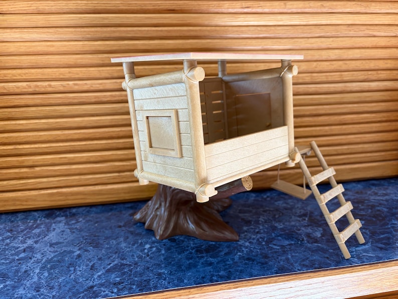 May include: A miniature wooden treehouse with a brown tree trunk base, a small ladder, and a swing. The treehouse has a light brown exterior with a square window and a flat roof. The background is a blue surface and a wooden wall.