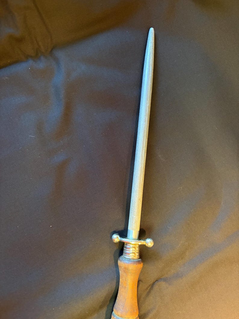 May include: A sharpening steel featuring a long, tapered, light gray metal rod with a textured surface. The tool includes a silver-colored metal guard with two rounded ends and a smooth, light brown wooden handle with a cylindrical shape.