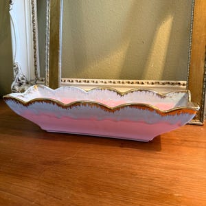 May include: A rectangular ceramic dish with a scalloped rim, featuring a smooth pink base that transitions into a white glaze toward the top edge. The scalloped edges are finished with a metallic gold trim, creating a distinct decorative border.