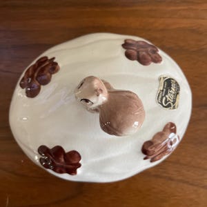 May include: White ceramic lid with a brown squirrel figurine on top. The lid is decorated with brown acorn designs and has a "Stardust" label.