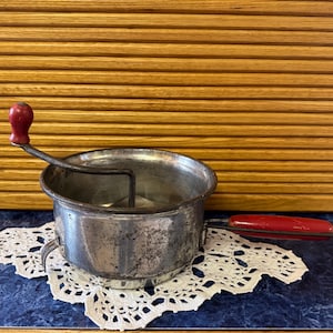 May include: A vintage metal food mill with a red handle and a red knob on the crank. The food mill sits on a white lace doily against a wood-paneled background. The food mill is a kitchen utensil.