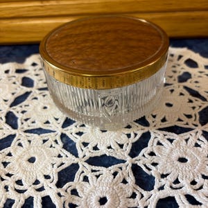 May include: A round glass trinket jar featuring vertical ribbed sides and a clear finish. The lid is circular with a textured, metallic gold-toned top surface and a smooth gold-colored rim that fits securely over the glass base of the container.