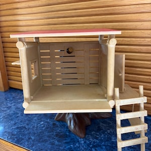 May include: A miniature wooden cabin with a red roof, a small window, and a ladder. The cabin is made of light brown wood and sits on a brown tree trunk base. The interior is visible, with horizontal wooden slats forming the walls.