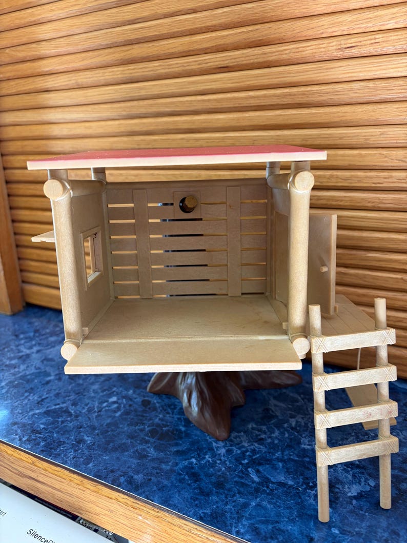 May include: A miniature wooden cabin with a red roof, a small window, and a ladder. The cabin is made of light brown wood and sits on a brown tree trunk base. The interior is visible, with horizontal wooden slats forming the walls.