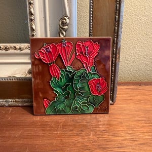 May include: Square ceramic tile featuring a botanical design. The tile showcases a cactus with vibrant red flowers and green leaves against a brown background. The artwork is detailed with fine lines and shading.