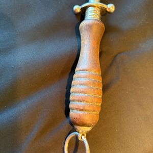 May include: A knife sharpening steel featuring a cylindrical metal rod, a polished silver-tone metal guard, and a contoured brown wooden handle with horizontal grooves. A circular metal hanging ring is attached to the base of the handle.
