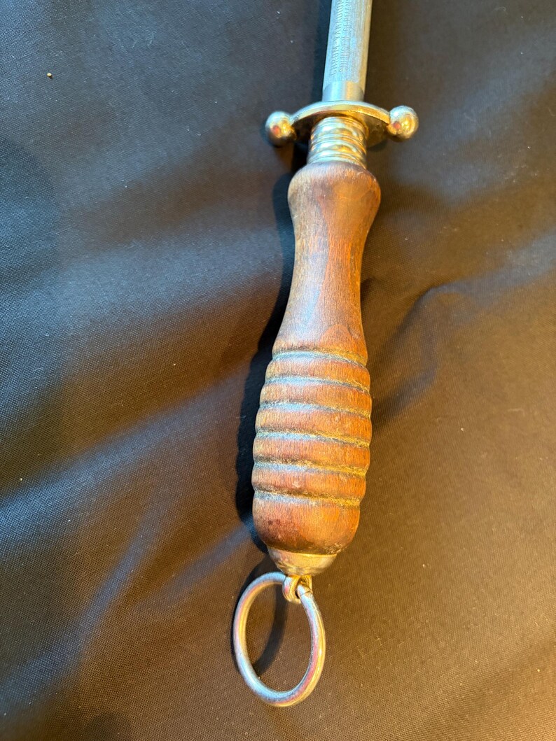 May include: A knife sharpening steel featuring a cylindrical metal rod, a polished silver-tone metal guard, and a contoured brown wooden handle with horizontal grooves. A circular metal hanging ring is attached to the base of the handle.