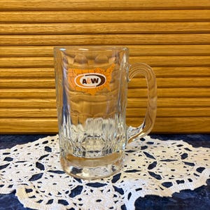 May include: Clear glass mug with a handle, featuring the A&W logo in orange and white. The mug has a textured design and sits on a white lace doily. The background includes a wooden surface.