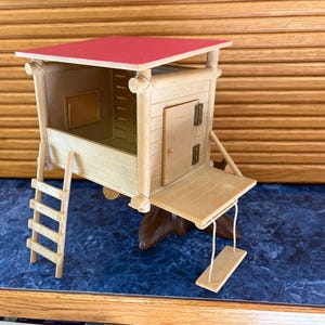May include: A miniature wooden treehouse with a red roof, a small door, and a ladder. The treehouse has a small swing hanging from the front. The toy is made of light-colored wood.