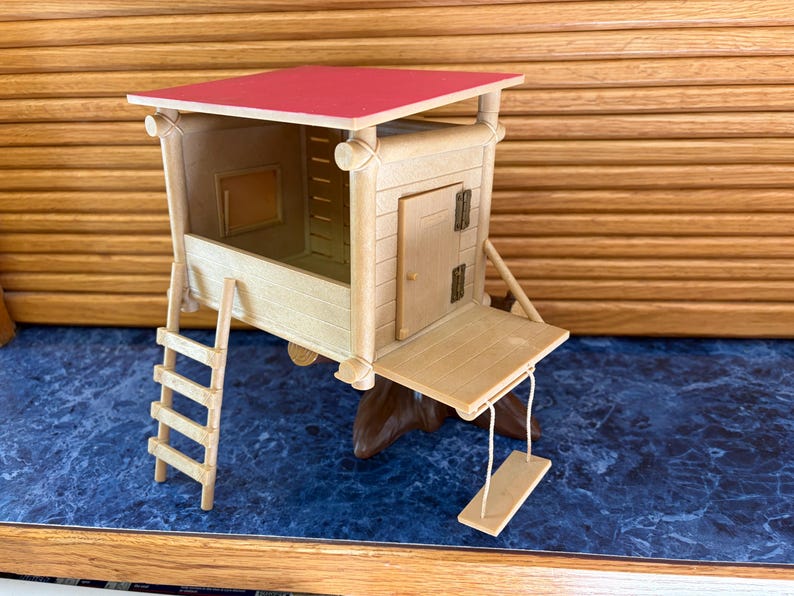 May include: A miniature wooden treehouse with a red roof, a small door, and a ladder. The treehouse has a small swing hanging from the front. The toy is made of light-colored wood.