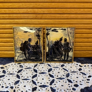 May include: Two framed silhouette art pieces depicting figures in a landscape. The black silhouettes are set against a light background, framed in gold. The artwork is displayed on a white lace doily, with a wooden background.