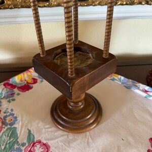 May include: A wooden stand with four vertical dowels and a square base. The stand has a turned pedestal base.