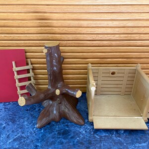 May include: A collection of toy components, including a brown tree trunk with branches, a tan wooden structure, a small wooden ladder, and a red rectangular piece. The items are arranged on a blue surface with a wooden slatted background.