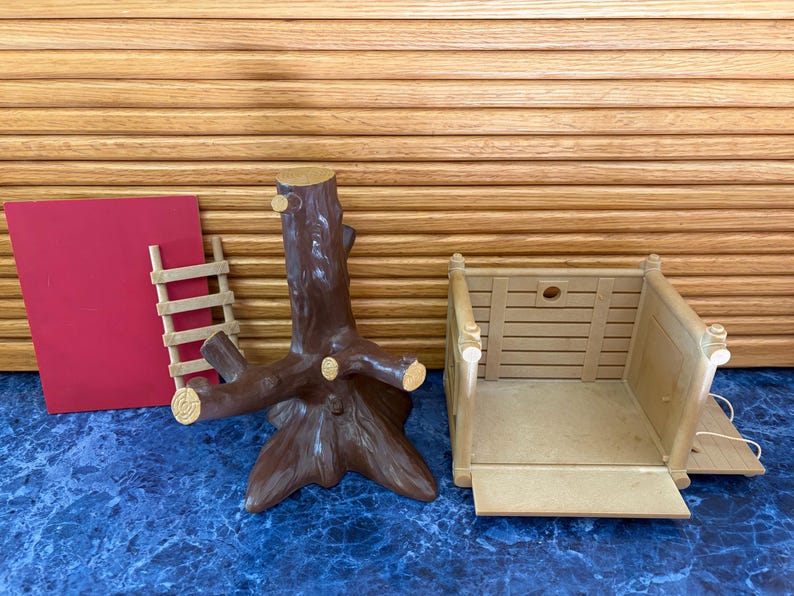 May include: A collection of toy components, including a brown tree trunk with branches, a tan wooden structure, a small wooden ladder, and a red rectangular piece. The items are arranged on a blue surface with a wooden slatted background.