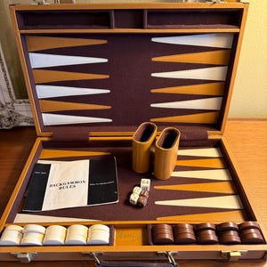 May include: A tan leather-bound backgammon set featuring a dark brown felt interior with white and tan triangular points. The set includes two tan dice cups, a set of white and brown checkers, four dice, a doubling cube, and a rule booklet.