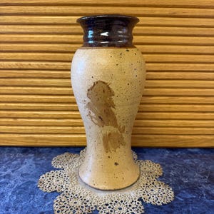 May include: A tan ceramic vase with a brown glaze around the top. The vase has a speckled brown design and is sitting on a white doily.