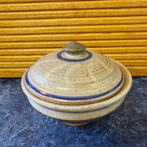 May include: A ceramic lidded bowl with a speckled, off-white glaze. The bowl and lid feature blue and brown bands. The lid has a small, rounded knob. The bowl is likely handmade and suitable for serving or storage.