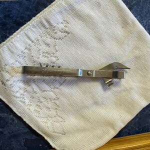 May include: A vintage, silver-colored combination can opener and bottle opener. The tool is resting on a white, patterned cloth. The can opener has a sharp blade and a lever for opening cans.