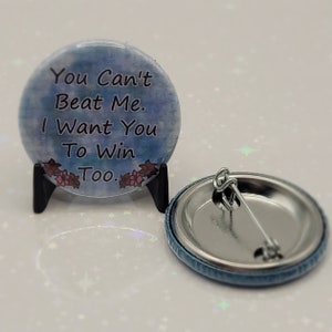 May include: A round button with the text "You Can't Beat Me. I Want You To Win Too." The button has a blue and purple watercolor background with floral accents. A second button is visible, showing the pin back.