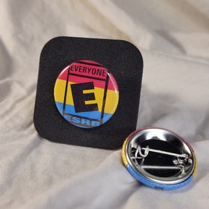 May include: A round button with a red, yellow, and blue background. The button has a black letter "E" in the centre and the text "EVERYONE" and "ESRB" in white. The button is on a black surface.