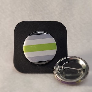 May include: A green, white and grey striped button badge with a silver pin back. The badge is on a black square background.