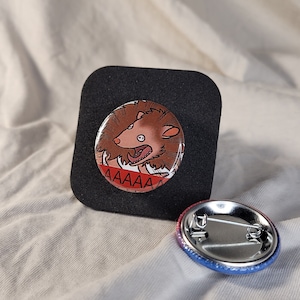 May include: A round button featuring a cartoon image of a brown and white hedgehog with a red mouth screaming "AAAAAAA". The button is displayed on a black backing.
