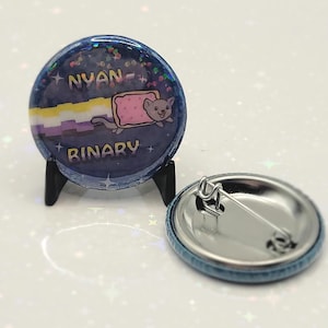 May include: A blue and purple button with a glittery finish features a cartoon cat wearing a pink and white bread slice. The text "NYAN - BINARY" is printed on the button.