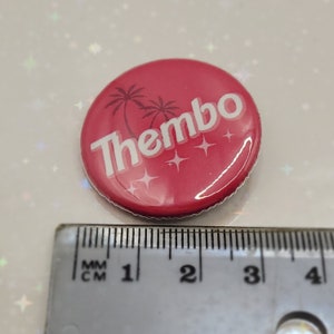 May include: A red button with a white design. The design features the word "Thembo" in a stylized font, two palm trees, and white stars. The button is approximately 1 inch in diameter.
