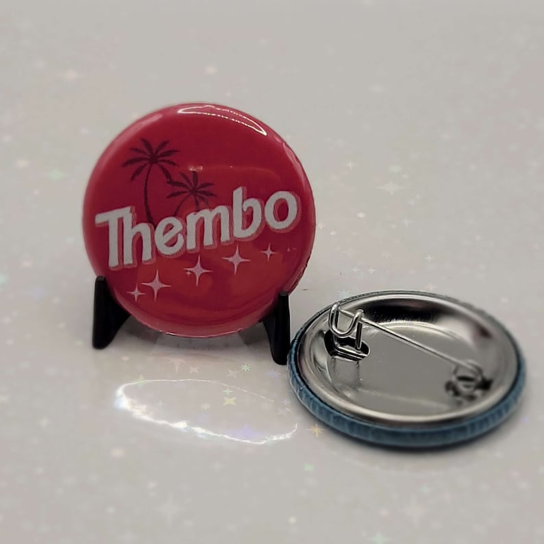 May include: A red button with a white palm tree design and the word "Thembo" in white lettering. The button has a silver back with a safety pin.
