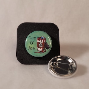 May include: A circular badge with a teal background and the words "Cup O' Joe". The badge features a cartoon coffee cup with an eyeball, blood, and a black object. A second badge with a pin back is also visible.
