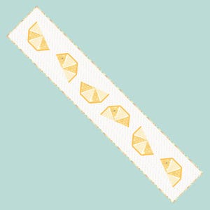 May include: A white rectangular table runner with a repeating pattern of stylized yellow lemon slices. The runner has a scalloped edge with a gold border, set against a light blue background.