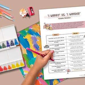 May include: A worksheet titled "I Worry vs. I Wonder" with examples, a watercolor palette, colored pencils, and a pencil sharpener. A hand holds a pink pencil, pointing to the worksheet. The worksheet is for self-reflection.