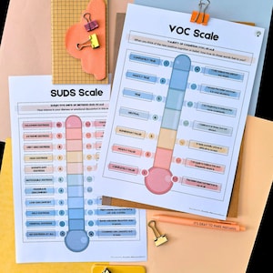May include: Two printed scales, the SUDS Scale and the VOC Scale, are displayed with a notebook, paper clips, and a pen. The SUDS scale measures distress levels, while the VOC scale assesses the validity of cognition. The pen has the text "IT'S OKAY TO MAKE MISTAKES".