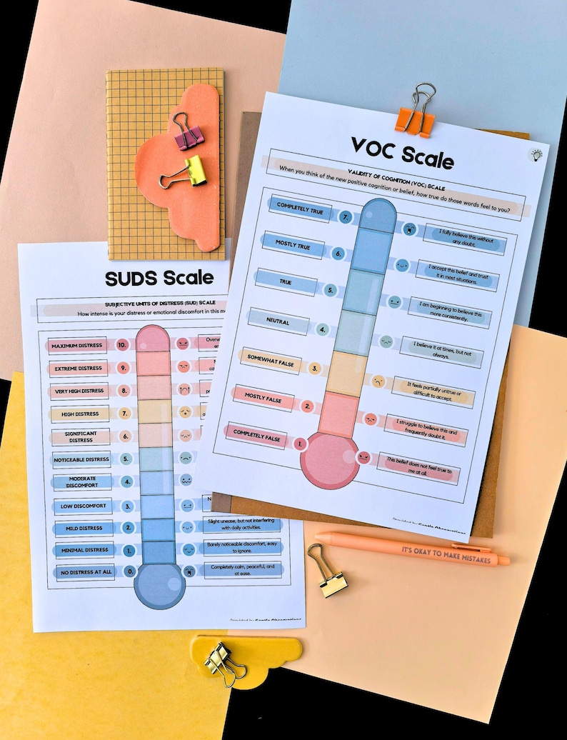 May include: Two printed scales, the SUDS Scale and the VOC Scale, are displayed with a notebook, paper clips, and a pen. The SUDS scale measures distress levels, while the VOC scale assesses the validity of cognition. The pen has the text "IT'S OKAY TO MAKE MISTAKES".