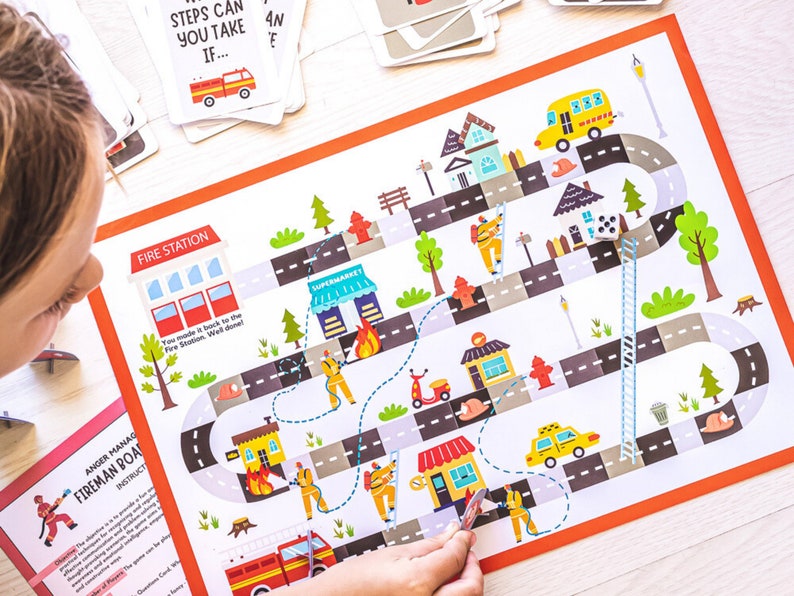 May include: A colorful board game with a fire station theme. The game board features a winding road with houses, a supermarket, a fire hydrant, and a fire truck. A child is playing the game with a die and a marker.